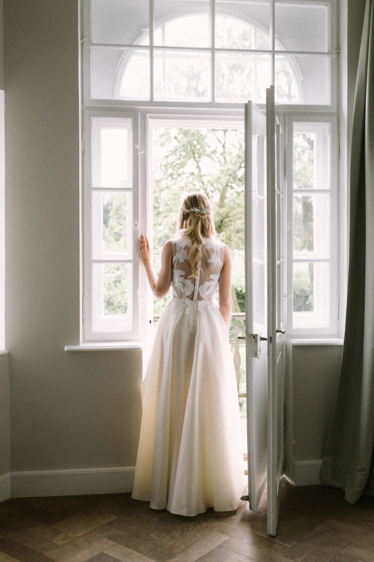 Die Braut fotografiert von hinten wie sie im Fenster steht, Hochzeit Gut Sonnenhausen - Alexandra Kasper